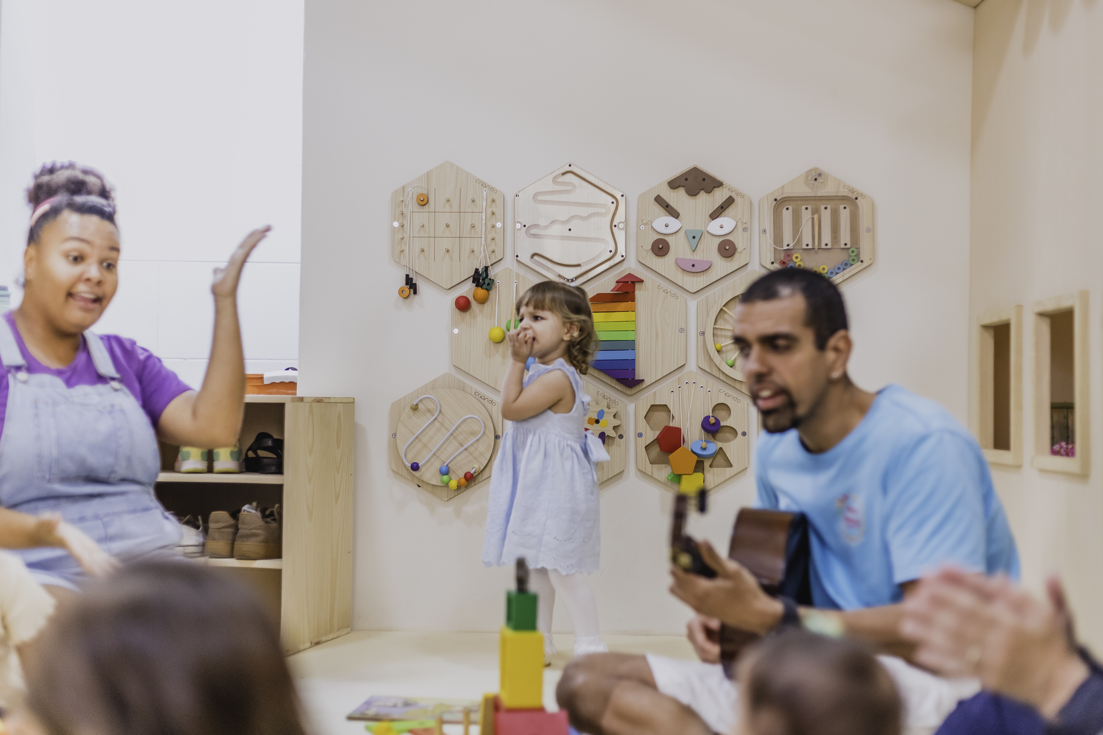 Monitor tocando violão ao lado de crianças brincando em painel de atividades