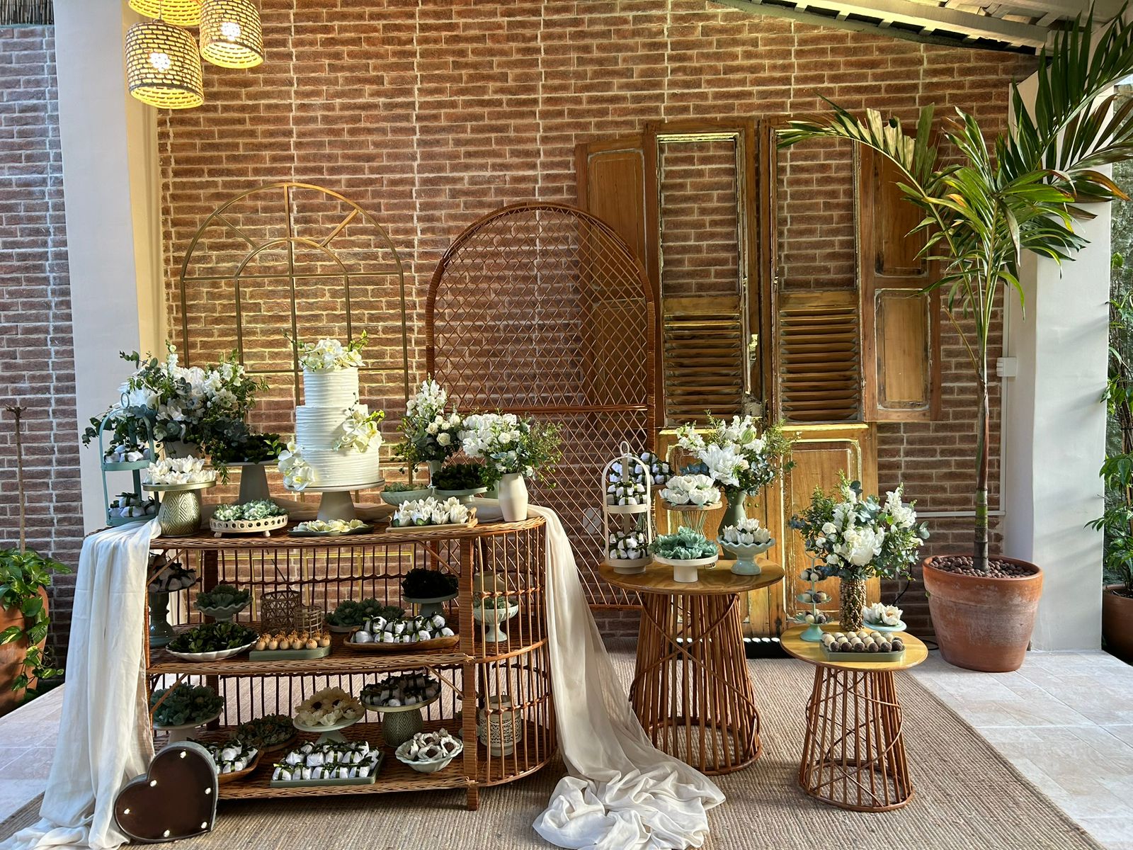 Mesa de doces branca com bolo de casamento em frente a parede de tijolinho