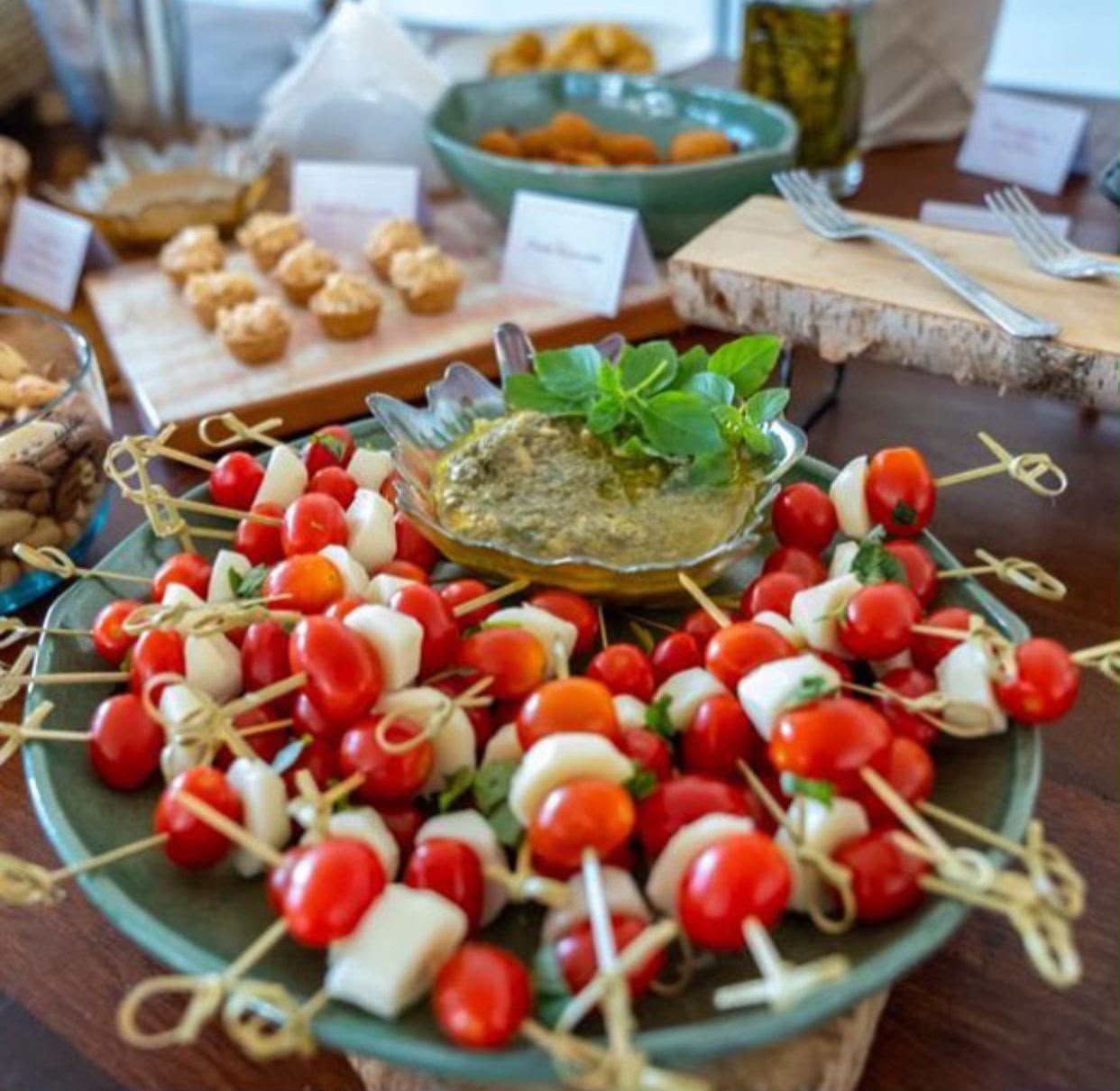 Espetinhos de tomate cereja e mussarela de búfala com molho pesto, mesa de buffet ao fundo